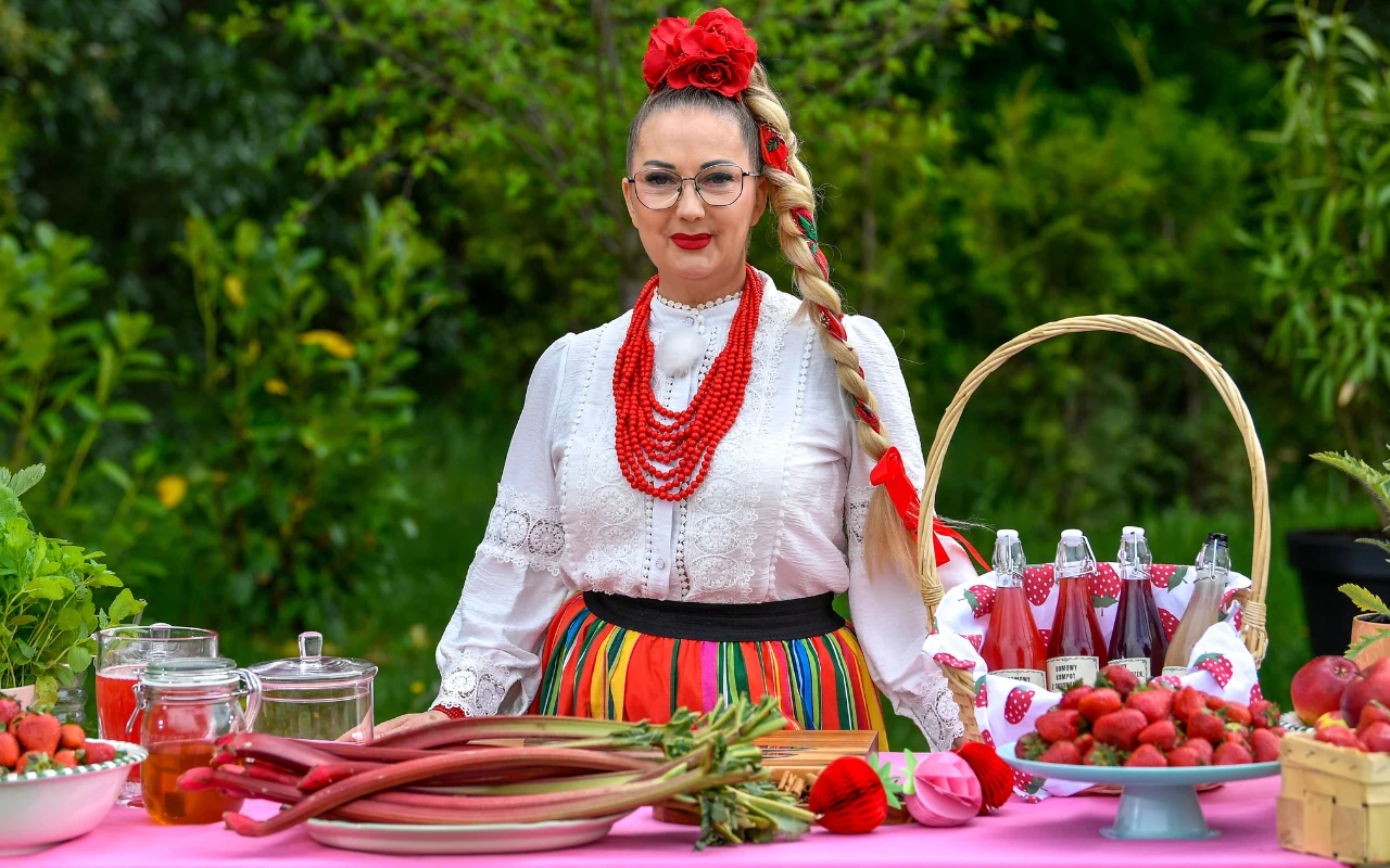 Gospodyni zdradziła sposób na idealny kompot. 3 proste zasady Magdalena Chomka w ludowym stroju za stołem z truskawkami i rabarbarem, zielone drzewa w tle