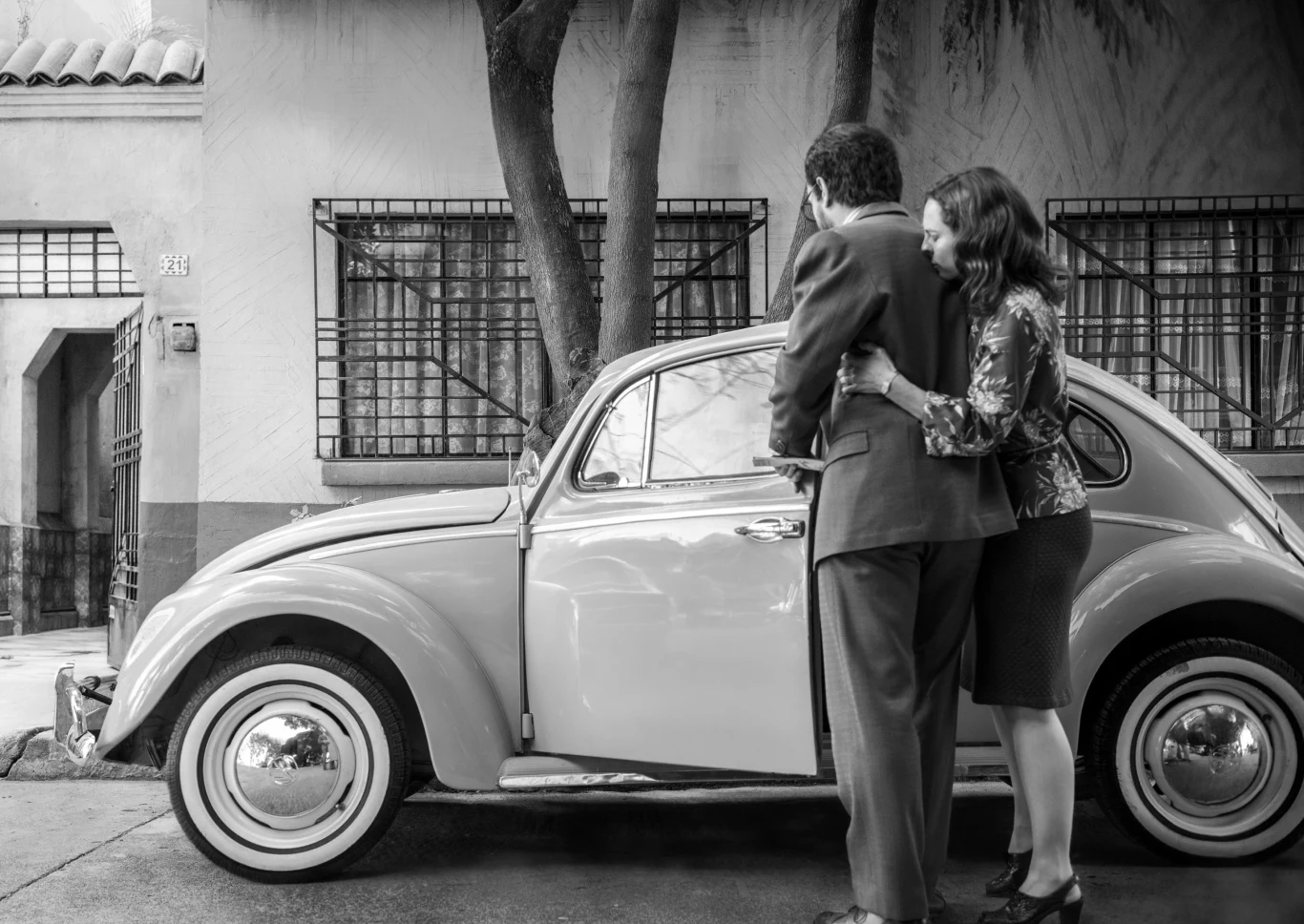 "Roma" Para elegancko ubranych osób stoi obok klasycznego samochodu typu garbus, w tle budynek mieszkalny z kratami w oknach.