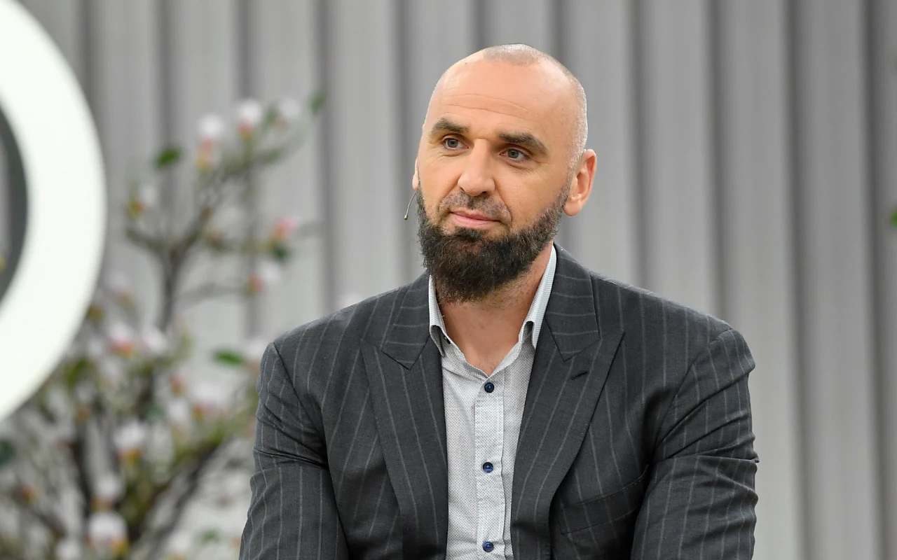 Brodaty mężczyzna o ogolonej głowie ubrany w szary garnitur i koszulę, siedzący na tle jasnych, pionowych paneli dekoracyjnych.