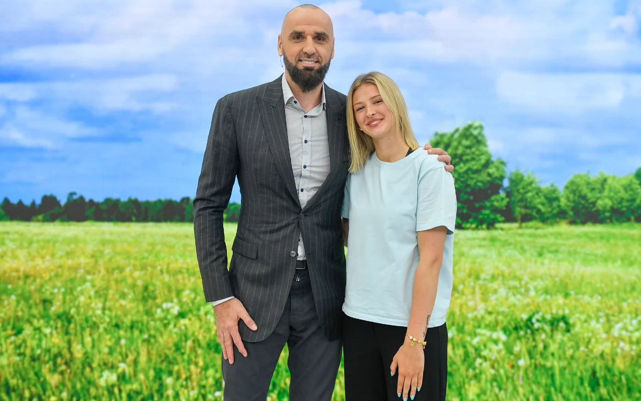 Mężczyzna w eleganckim garniturze obejmuje uśmiechniętą kobietę w jasnej koszulce, stojąc razem na tle zielonej łąki i niebieskiego nieba.