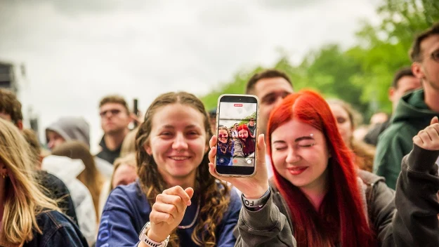 Dwie roześmiane młode osoby stoją przy metalowej barierce na tle tłumu, jedna z nich trzyma telefon pokazujący selfie z uśmiechem obojga.