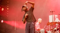 Dwóch muzyków podczas występu scenicznego – wokalista w czarnej koszulce gestykuluje na pierwszym planie, a w tle perkusista gra na bębnach, obaj oświetleni czerwonym światłem scenicznym.
