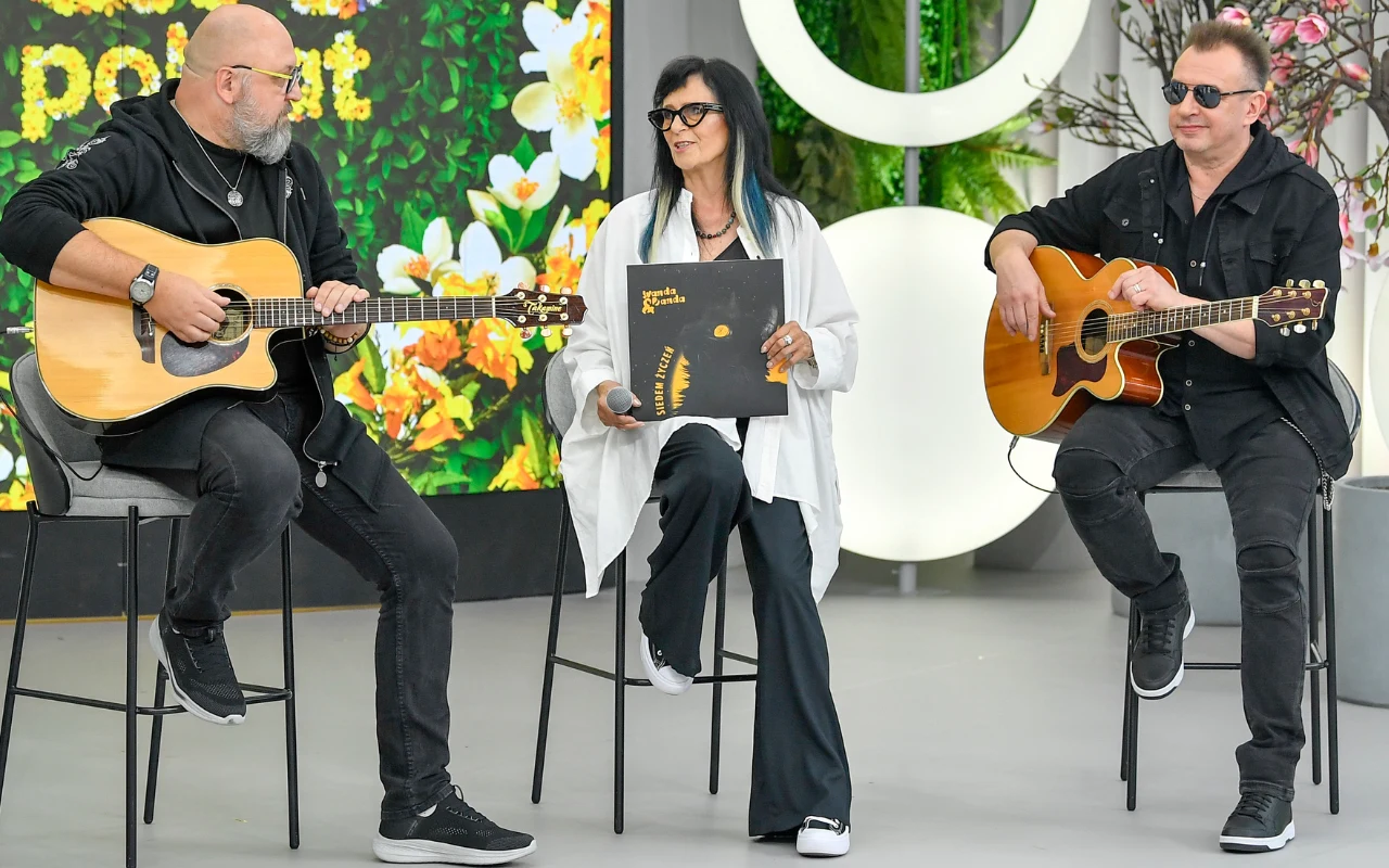 Dwóch mężczyzn grających na gitarach akustycznych siedzi po obu stronach kobiety, która trzyma dużą czarną płytę winylową z żółtymi napisami.