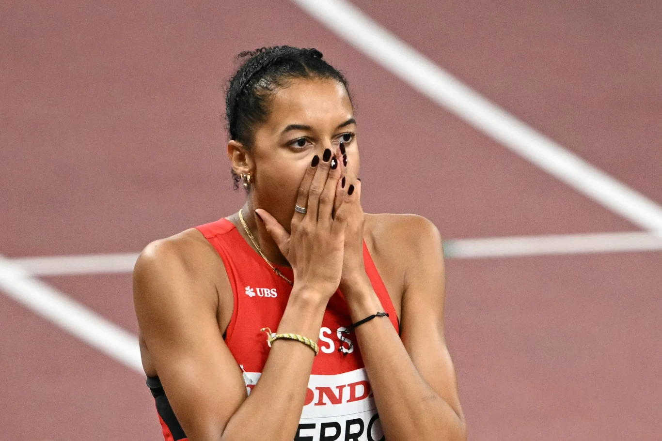 Zawodniczka w czerwonym stroju sportowym z logo sponsoringu zakrywa usta dłońmi, prezentując silne emocje po zakończeniu biegu na stadionie lekkoatletycznym.