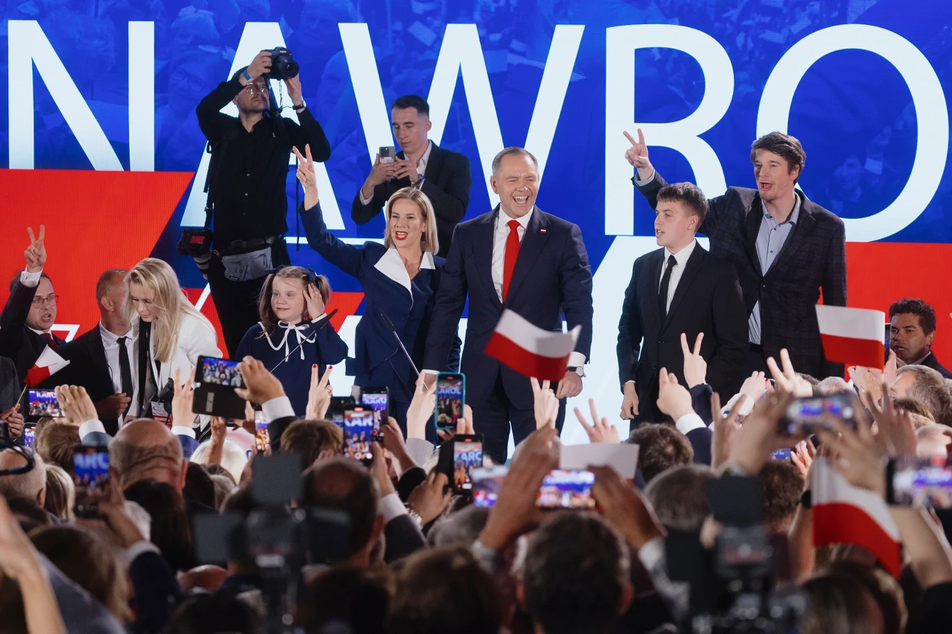 Uśmiechnięty mężczyzna w garniturze stoi na scenie wśród kilku osób, trzymając flagę Polski, otoczony tłumem ludzi z telefonami i flagami, na tle dużego napisu z biało-czerwonymi elementami.