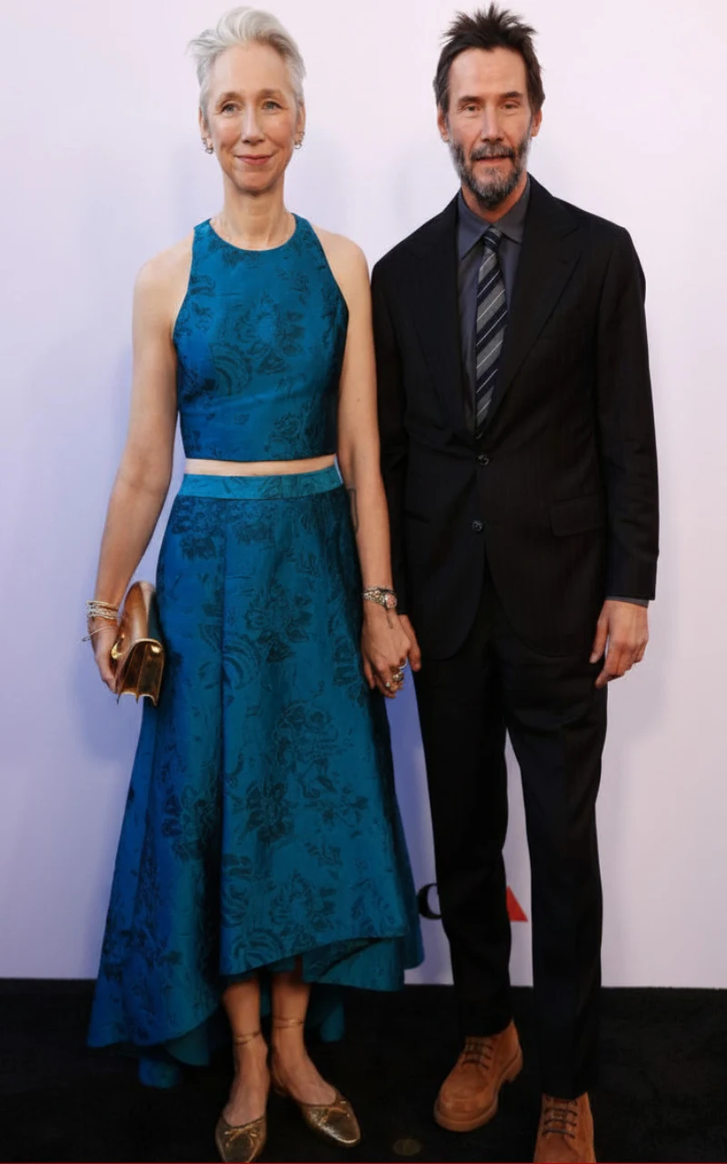 Alexandra Grant i Keanu Reeves Kobieta w eleganckiej, niebieskiej sukni z połyskiem oraz mężczyzna w ciemnym garniturze stoją obok siebie, trzymając się za ręce; kobieta trzyma złotą kopertówkę.