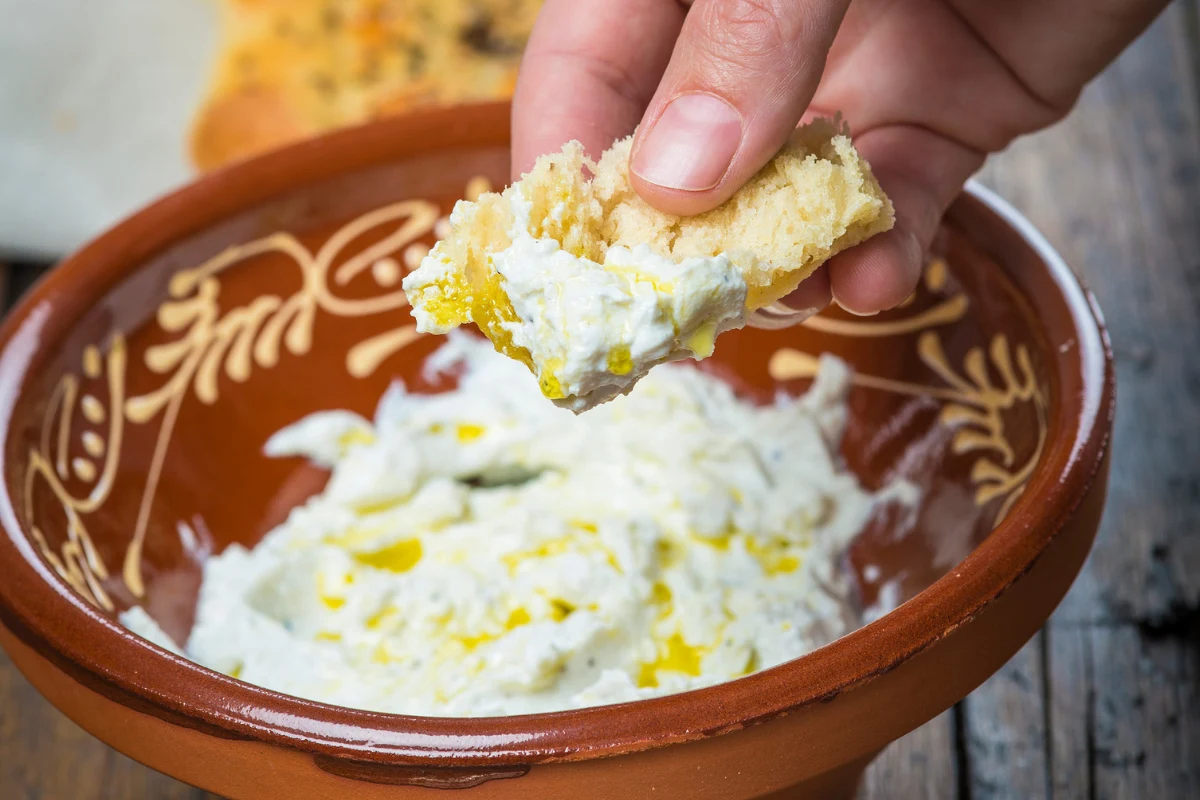Labneh na kanapki - jakie ma właściwości? Chleb maczany w labneh z oliwą w ceramicznej misce, ujęcie dłoni trzymającej kromkę.