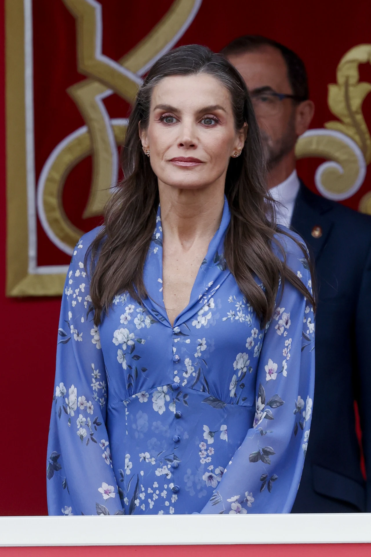Królowa jest wierna hiszpańskiej modzie i przy kluczowych wydarzeniach jej wybór pada właśnie na hiszpańskich projektantów Kobieta w eleganckiej, niebieskiej sukience w kwiaty stoi na tle czerwono-złotej dekoracji, z poważnym wyrazem twarzy, z rozpuszczonymi włosami, patrząc przed siebie.