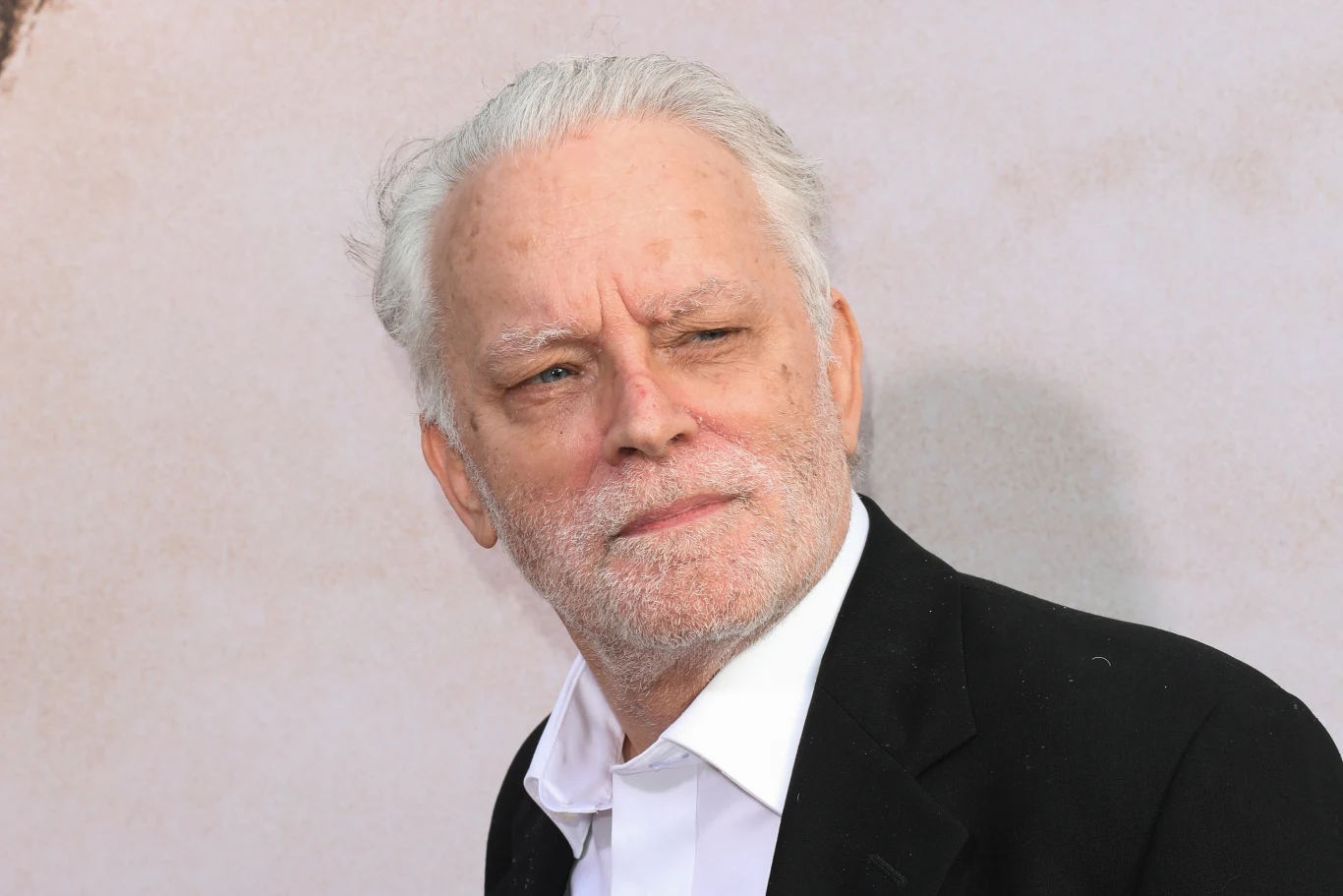 Brad Dourif w 2019 roku Starszy mężczyzna z siwymi włosami i zarostem, ubrany w czarną marynarkę i białą koszulę, patrzący lekko w bok na tle jasnej ściany.