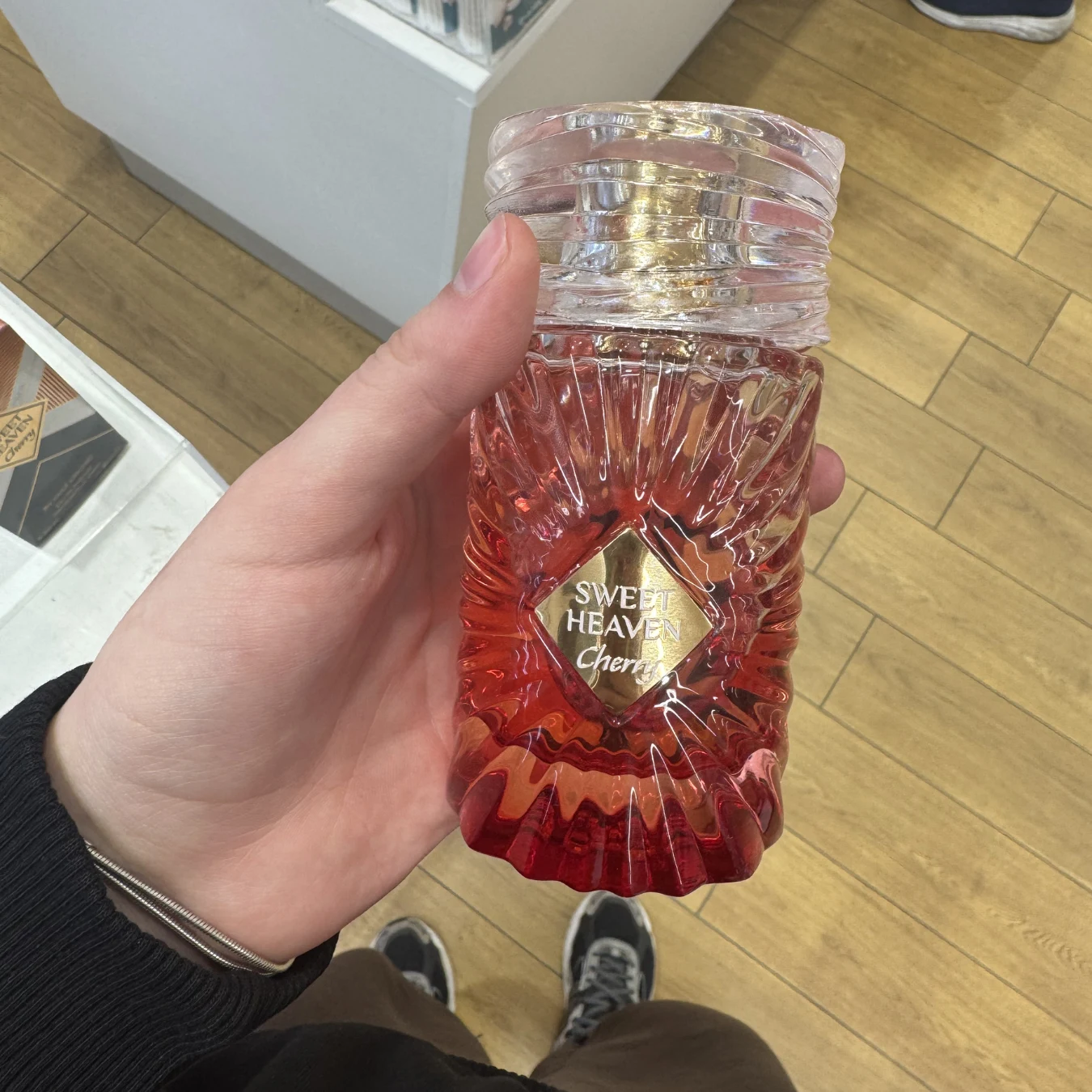 Ręka trzyma szklaną butelkę perfum o nazwie Sweet Heaven Cherry w czerwono-różowym kolorze z tłoczeniami, w tle jasna podłoga i fragment sklepowej lady.