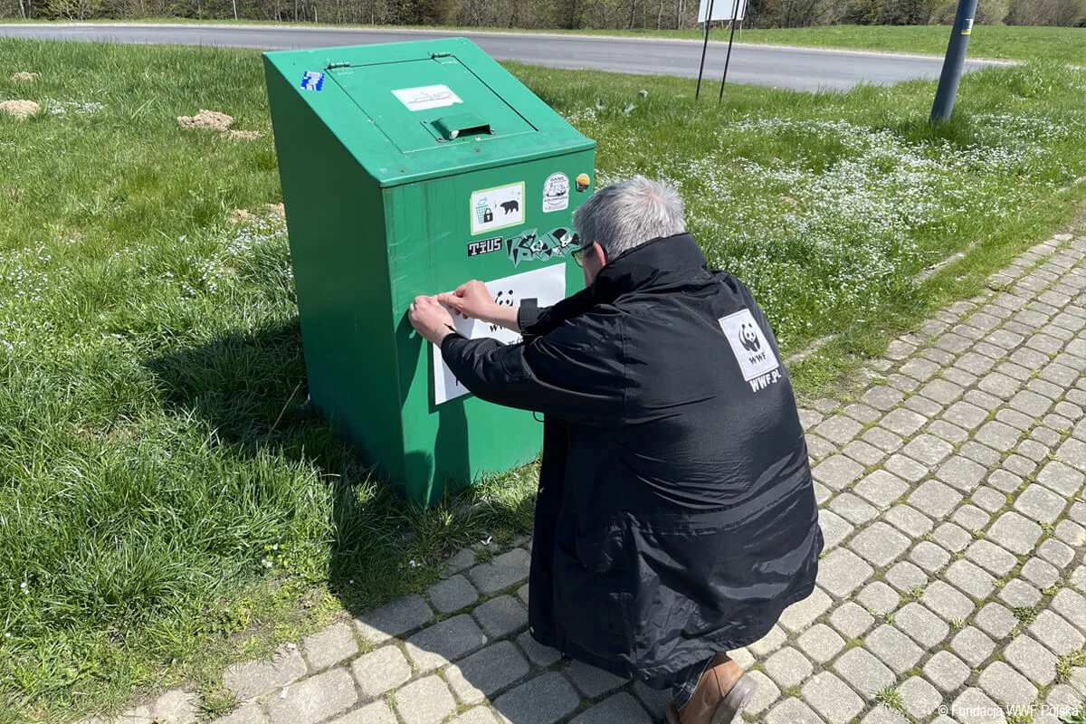 Osoba w czarnym płaszczu z logo WWF przykleja plakat na zielonym kontenerze stojącym na trawie, obok brukowanego chodnika.