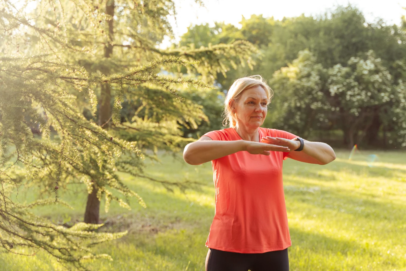 Kobieta w średnim wieku w sportowej koszulce wykonuje ćwiczenia na świeżym powietrzu, otoczona zielenią drzew i trawy.