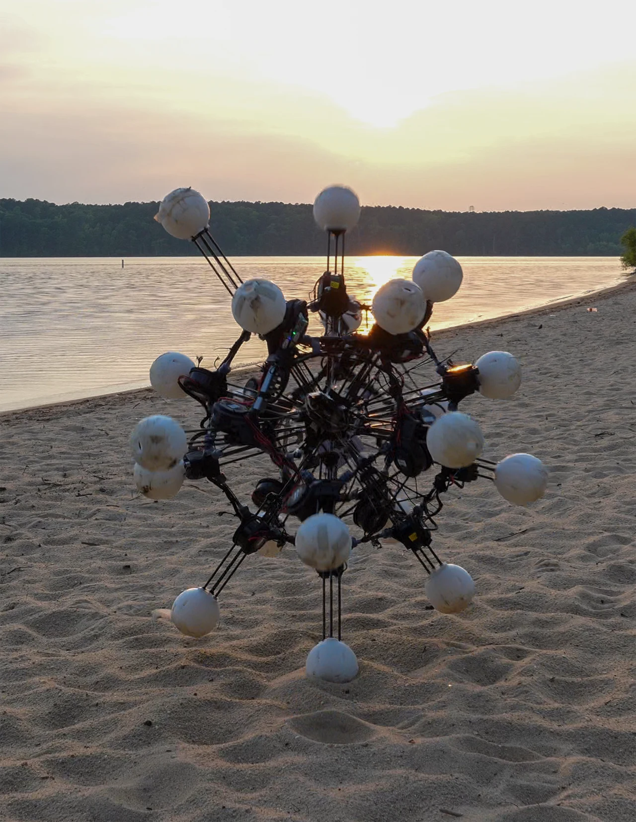 Futurystyczny, wieloramienny robot z kulistymi końcówkami porusza się po piaszczystej plaży nad jeziorem o zachodzie słońca.