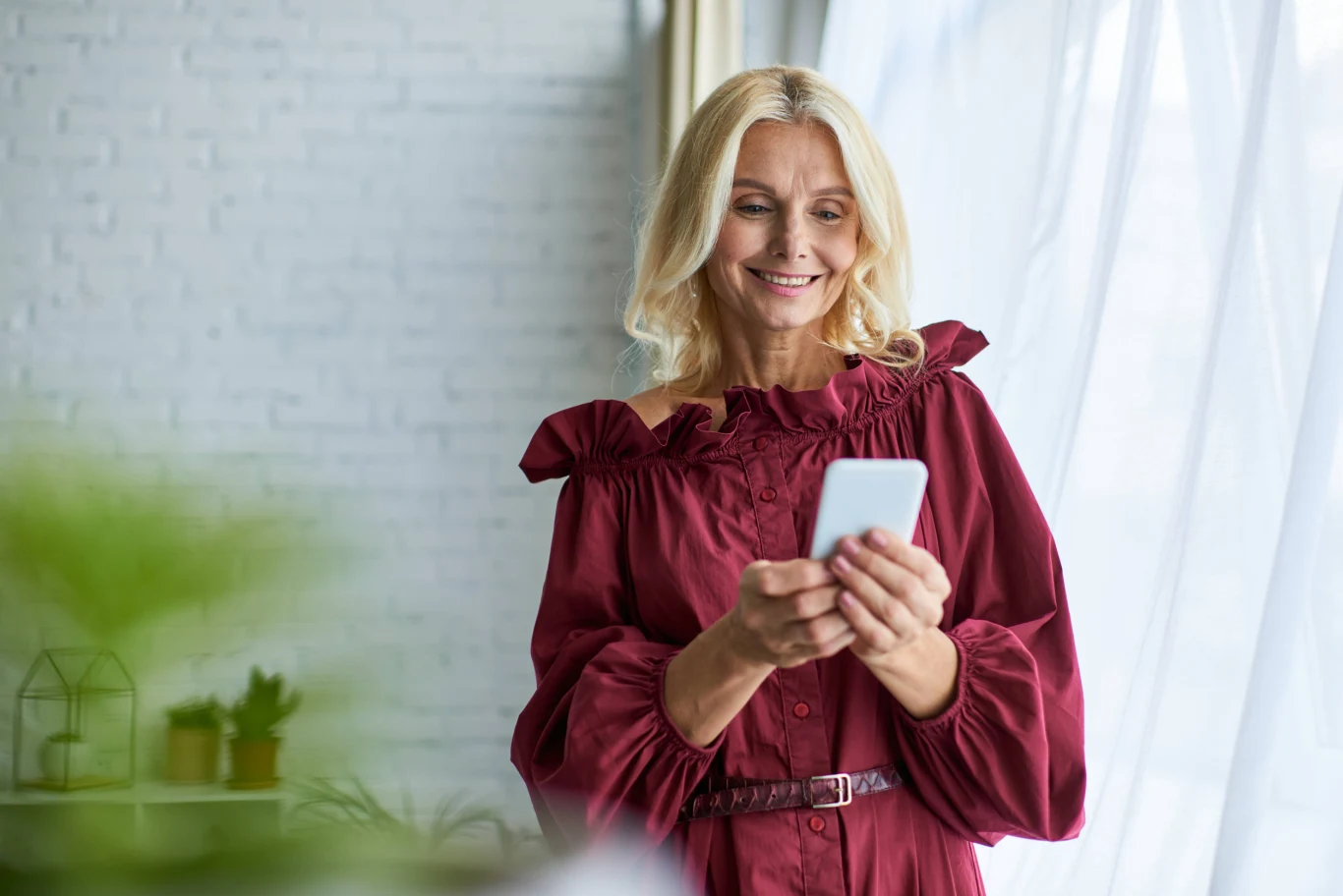 Uśmiechnięta kobieta w bordowej sukience trzyma w dłoniach smartfon i patrzy na ekran, stojąc przy jasnym oknie.