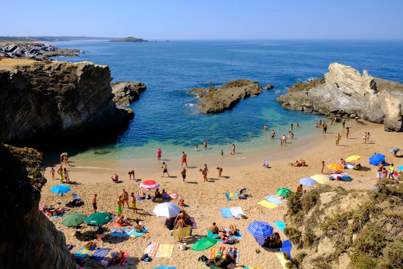 Tłum ludzi wypoczywających na piaszczystej plaży otoczonej skalistymi klifami, kolorowe parasole, kąpiący się w spokojnej, błękitnej wodzie oraz opalający się na piasku urlopowicze.