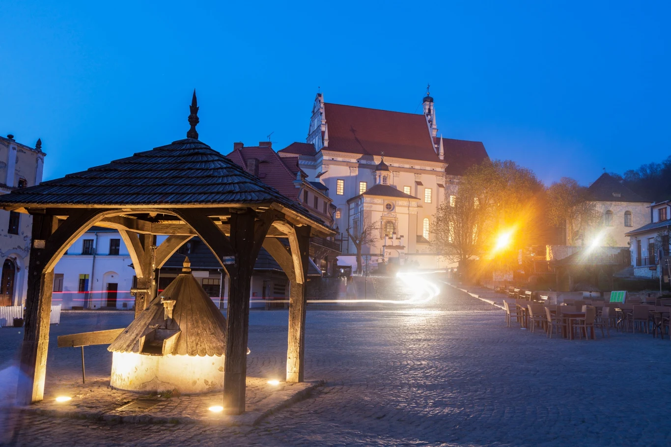 Stara studnia na rynku w Kazimierzu Dolnym Zabytkowa studnia z drewnianym zadaszeniem stojąca na rynku otoczona kamienicami, w tle oświetlony kościół z charakterystycznym czerwonym dachem.