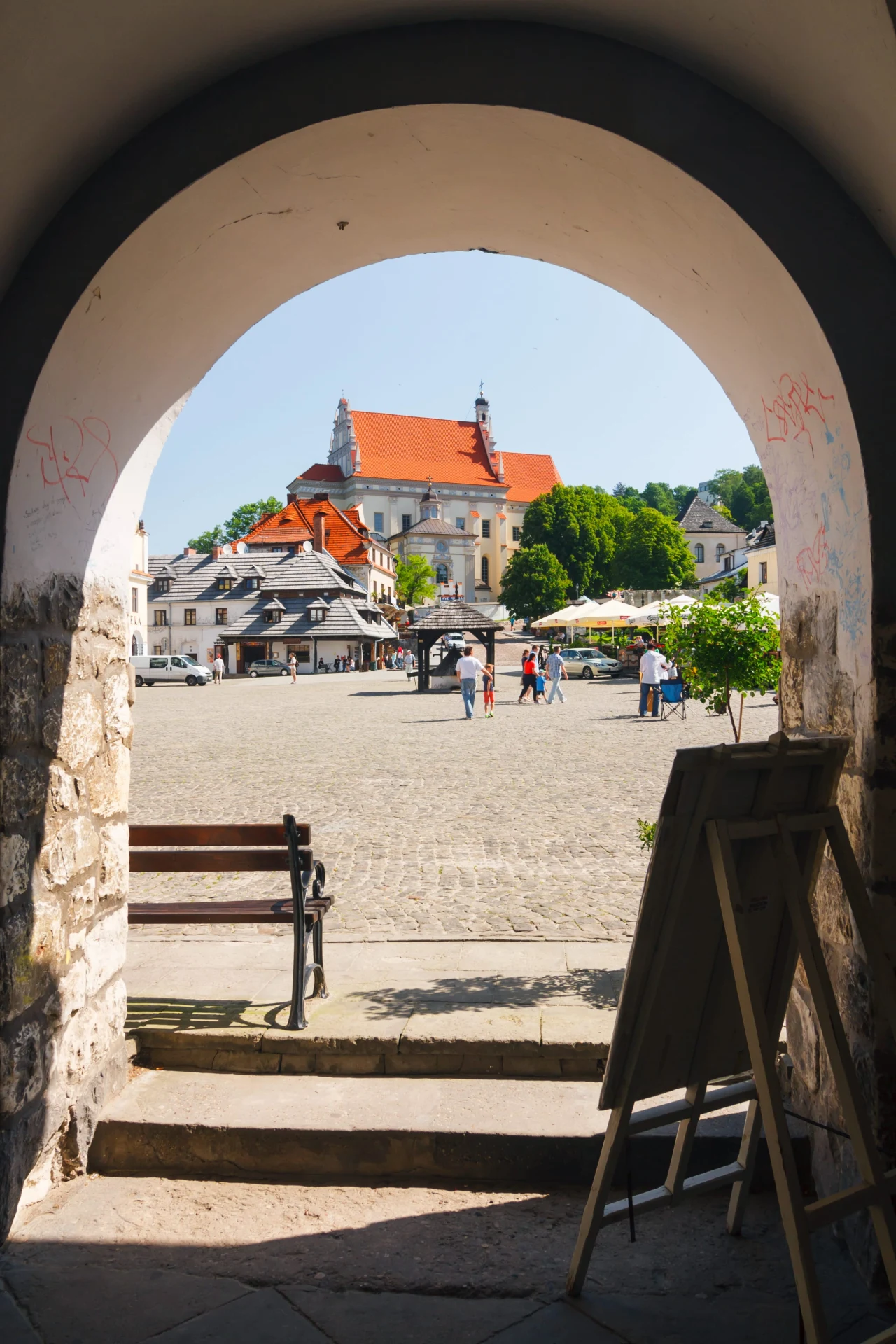 Kazimierz Dolny jest bardzo klimatyczny Kamienny łuk bramy prowadzący na główny rynek z brukowaną nawierzchnią, w tle kościół z czerwonym dachem, otoczony zielenią oraz kilkoma spacerującymi osobami.