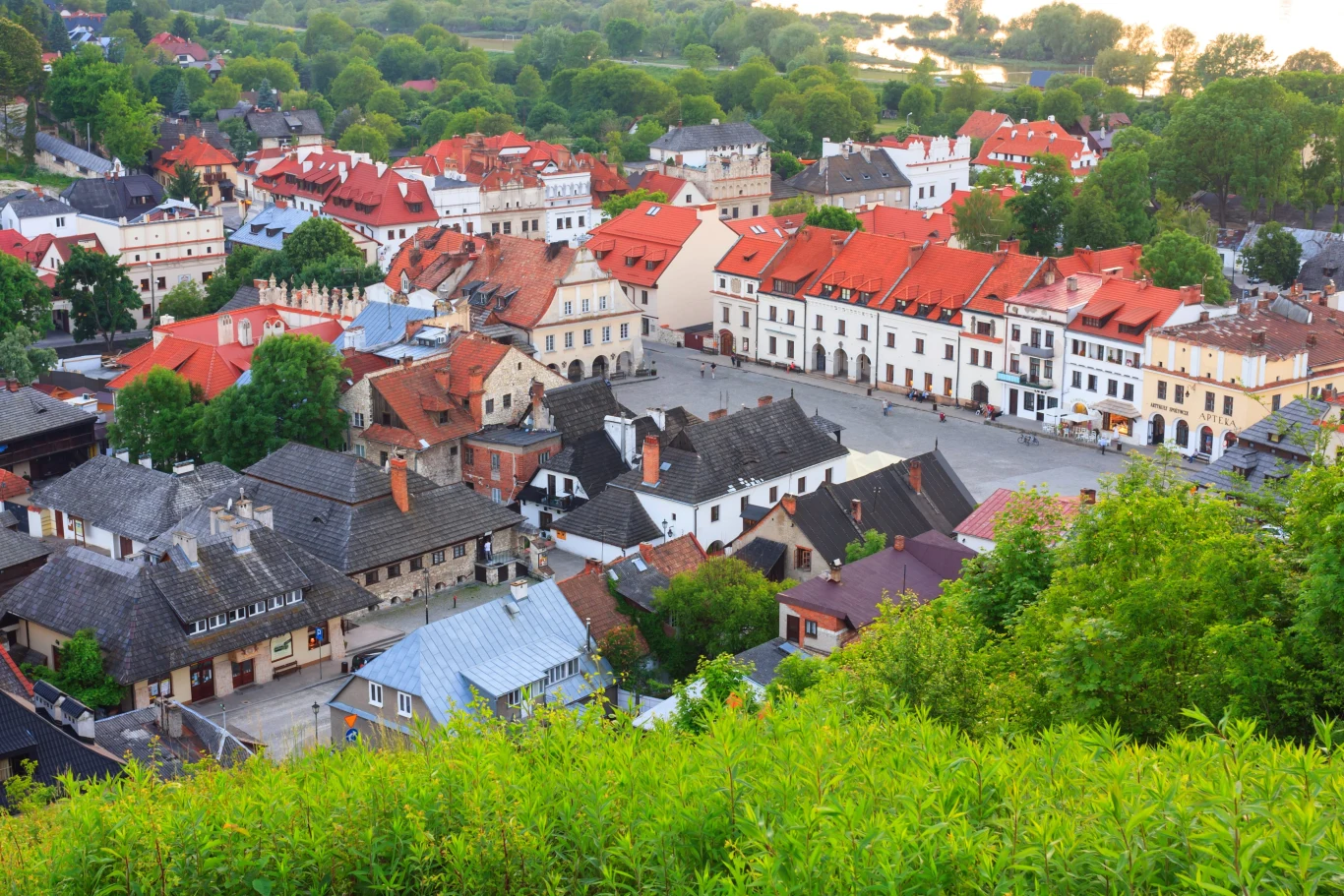 Widok z lotu ptaka na Kazimierz Dolny Rynek otoczony zabytkowymi kamienicami z czerwonymi dachami, pośród zieleni i w otoczeniu malowniczych, historycznych budynków.