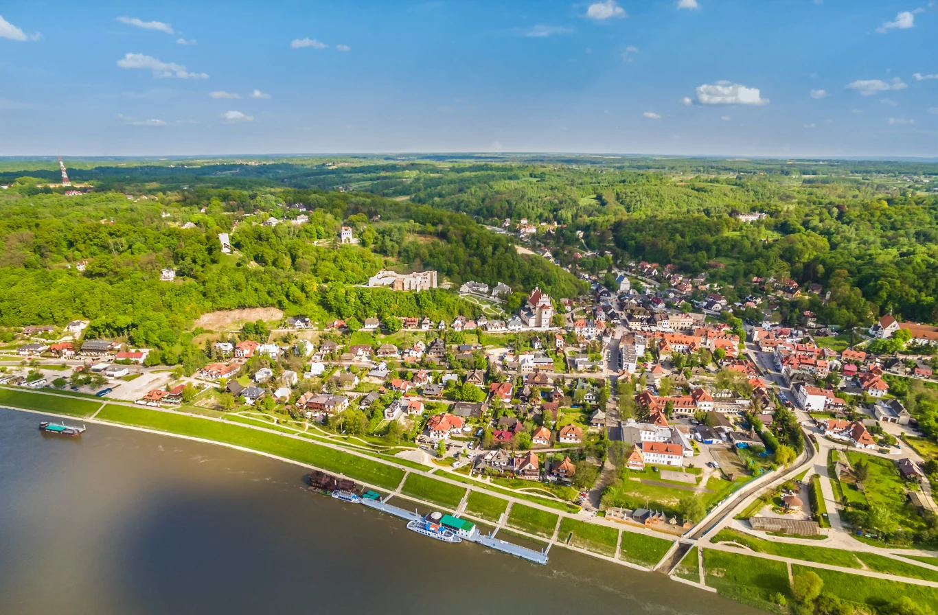 Kazimierz Dolny z lotu ptaka Panorama niewielkiego miasteczka położonego nad rzeką z portem i licznymi domami otoczonymi zielonymi wzgórzami i lasami.