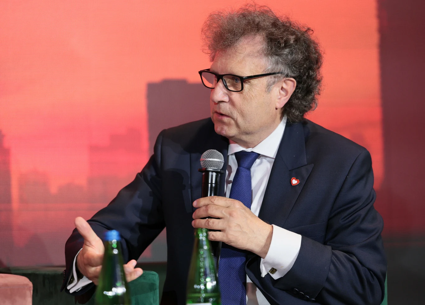 Jacek Karnowski: Kolejna transza pieniędzy z KPO może trafić do Polski w przyszłym tygodniu Jacek Karnowski w okularach i garniturze z przypinką, mówi do mikrofonu, na stole dwie butelki wody.