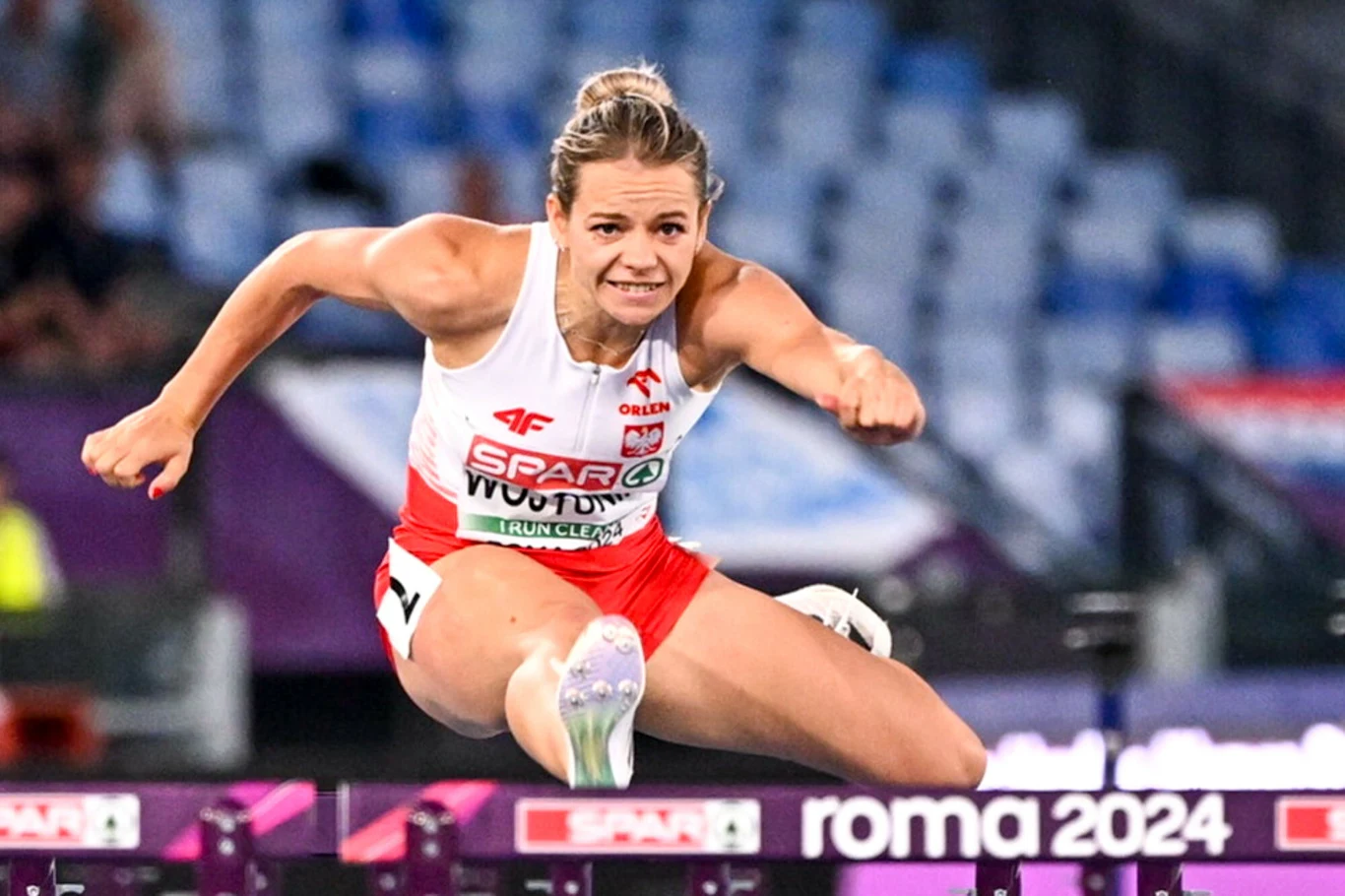 Lekkoatletka w biało-czerwonym stroju przeskakująca płotek podczas biegu na stadionie, widoczne oznaczenie zawodów Roma 2024.