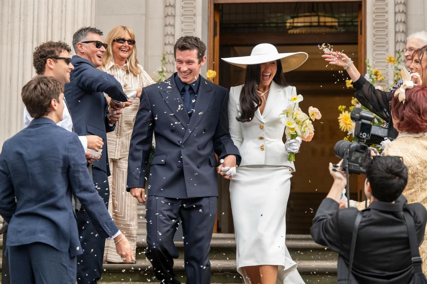 Para ubrana elegancko, wychodząca z budynku po schodach, uśmiechnięci goście otaczają ich i rzucają konfetti, w tle widać aparat fotograficzny i kwiaty w żółtych odcieniach.