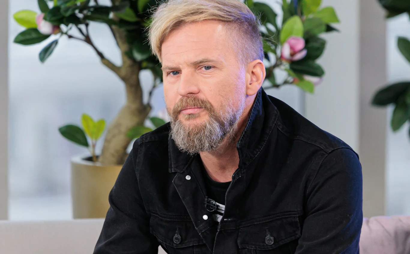 Tomasz Organek Tomasz Organek z blond brodą ubrany w czarną kurtkę jeansową siedzi na jasnej sofie.