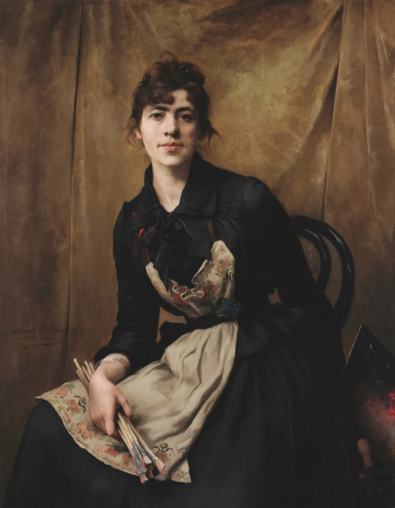 Anna Bilińska-Bohdanowiczowa, autoportret z 1887 roku Młoda kobieta z jasną cerą i upiętymi włosami siedząca na czarnym krześle, ubrana w ciemną suknię z jasnym fartuchem, trzyma kilka pędzli malarskich w jednej dłoni oraz paletę w drugiej.