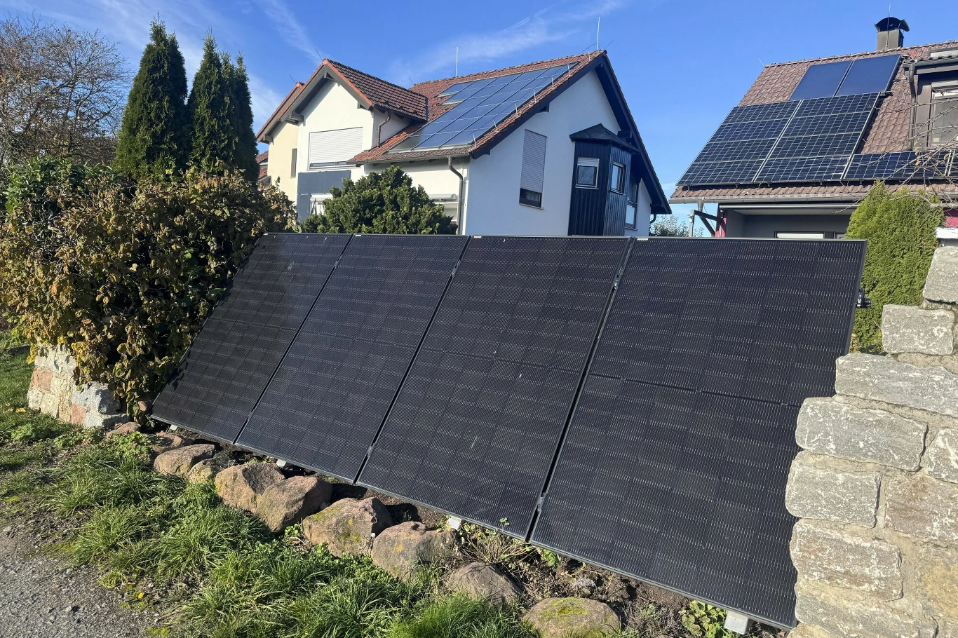 Panele fotowoltaiczne ustawione na trawniku przed domami, widoczne także instalacje solarne na dachach budynków.