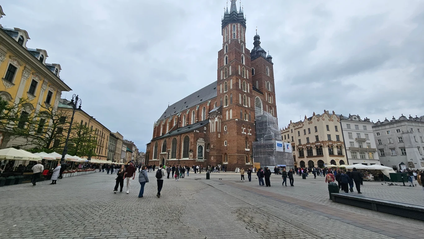 Kościół Mariacki górujący nad Rynkiem Głównym w Krakowie, liczni przechodnie oraz kawiarniane ogródki po lewej stronie placu.