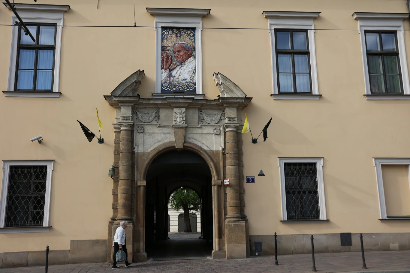 Historyczna brama z łukiem zdobiona portretem Jana Pawła II umieszczonym nad wejściem, przy której powiewają czarne i żółte flagi, a obok przechodzi samotna osoba.
