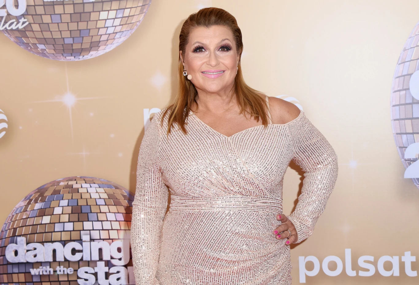 Kobieta w połyskującej, jasnej sukni z długim rękawem pozuje na tle scenograficznym programu Dancing with the Stars, w otoczeniu błyszczących kul dyskotekowych i logo Polsatu.