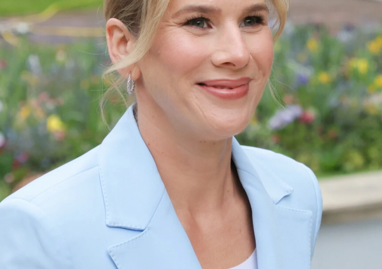 Marta Nawrocka Marta Nawrocka w jasnobłękitnej marynarce i białym topie, z upiętymi blond włosami, na tle kwiatów.