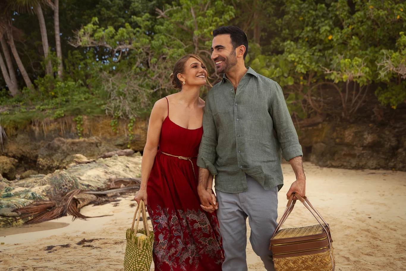 Jennifer Lopez i Brett Goldstein w filmie "Biurowy romans" Para trzymająca się za ręce spaceruje po piaszczystej plaży, niosąc piknikowe kosze, uśmiechnięci i otoczeni drzewami.