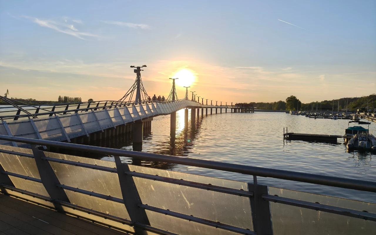 Most pieszy rozciągający się nad spokojną taflą jeziora, oświetlony promieniami zachodzącego słońca, z łodziami zacumowanymi przy pomostach po prawej stronie.