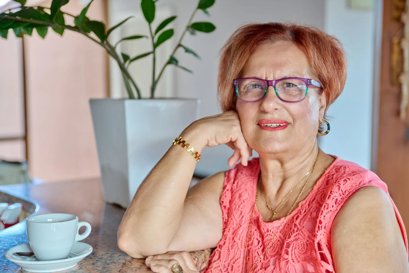 Starsza kobieta w okularach i koralowej bluzce siedzi przy stole z filiżanką kawy, w tle duża doniczka z zieloną rośliną.