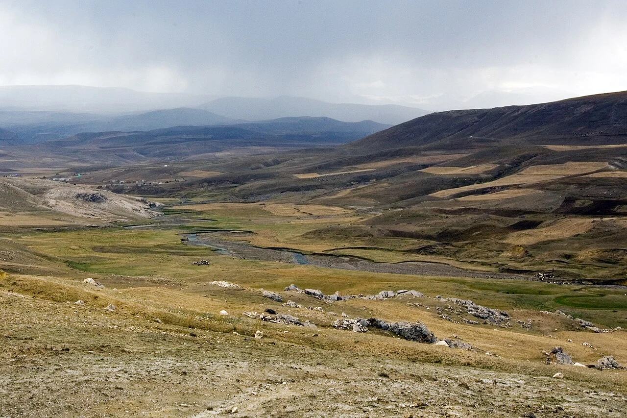 Rozległe, pagórkowate tereny porośnięte suchą trawą i przetykane niewielkimi skałami, przez które meandruje wąska rzeka pod pochmurnym niebem.