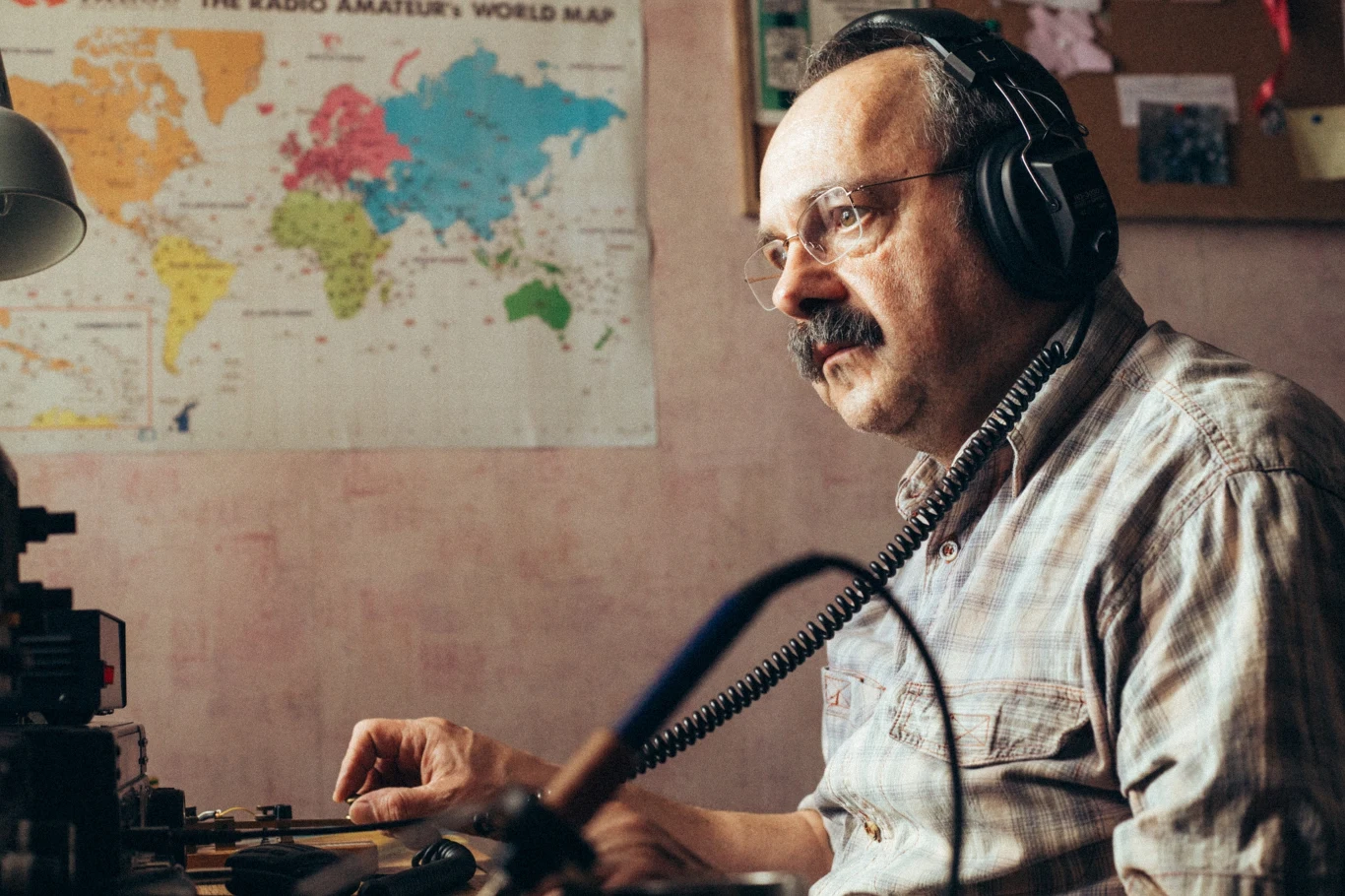 Mężczyzna w słuchawkach siedzący przy radiostacji, z mapą świata na ścianie w tle.