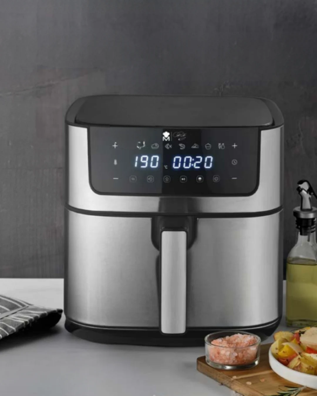 Srebrna frytkownica beztłuszczowa z cyfrowym panelem dotykowym ustawionym na 190°C i 20 minut, obok butelka z oliwą, rękawica kuchenna oraz miseczki z przygotowanymi składnikami do gotowania.
