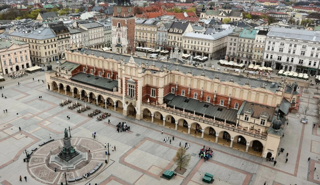 Sukiennice w centrum krakowskiego Rynku Głównego z widoczną wieżą Kościoła Mariackiego w tle, wokół rozległy brukowany plac z pomnikiem Adama Mickiewicza i układem zabytkowych kamienic.