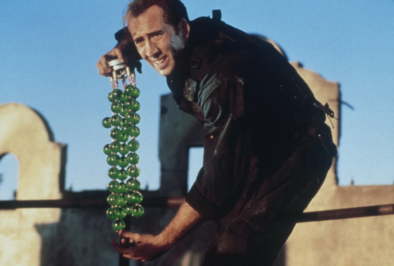 Nicolas Cage w filmie "Twierdza" Mężczyzna w czarnym kombinezonie taktycznym trzyma długi pręt z połączonymi zielonymi kulami, skoncentrowany na ostrożnym manewrowaniu w niebezpiecznej sytuacji.