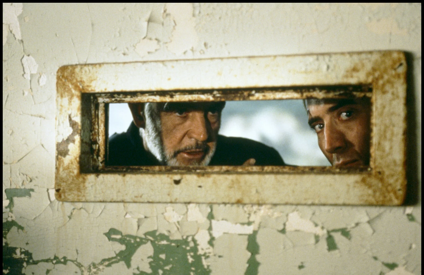 Sean Connery i Nicolas Cage w filmie "Twierdza" Dwie twarze mężczyzn spoglądające przez prostokątny otwór w zniszczonych drzwiach z odpryskującą farbą.