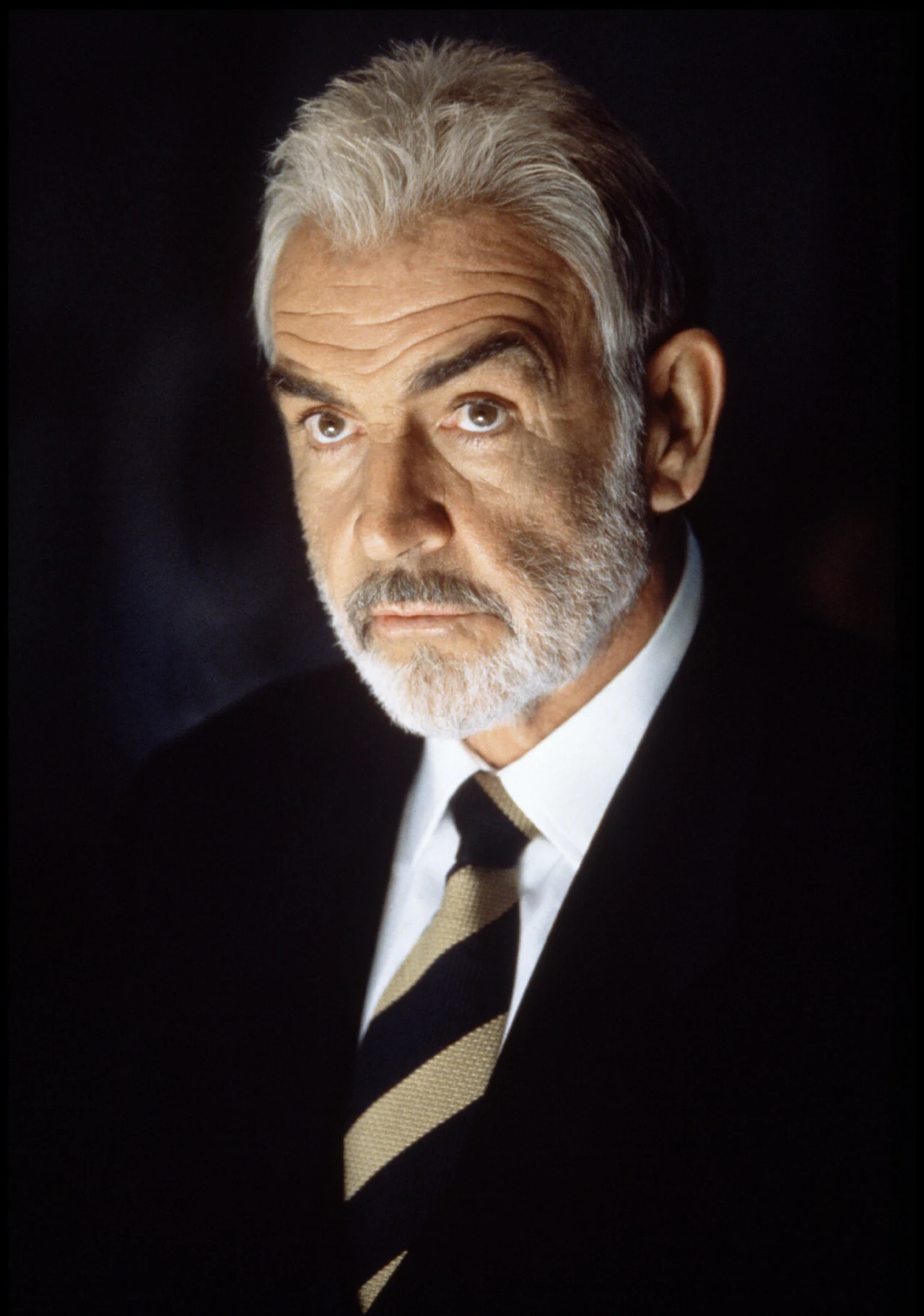 Sean Connery w filmie "Twierdza" Mężczyzna o siwych włosach i brodzie, ubrany w ciemny garnitur, białą koszulę i krawat w szerokie, ukośne pasy, spoglądający poważnie w stronę obiektywu.