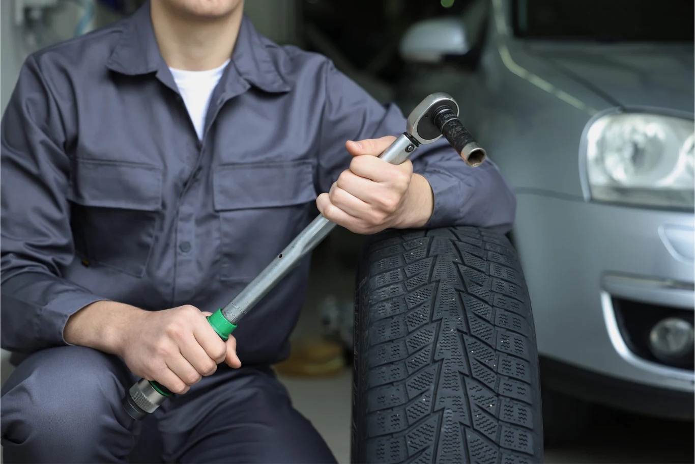 Mechanik w roboczym kombinezonie trzyma klucz dynamometryczny i opiera się o oponę obok samochodu osobowego w warsztacie.