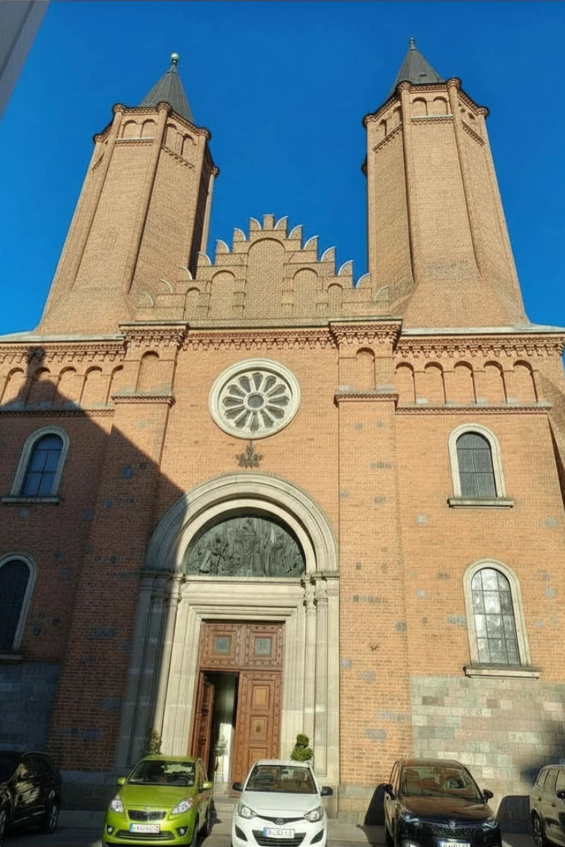 Ceglana fasada kościoła z dwiema wysokimi wieżami, centralnym dużym portalem wejściowym otoczonym kamiennym obramowaniem oraz dekoracyjnym okrągłym witrażem nad drzwiami, przed budynkiem zaparkowane samochody.