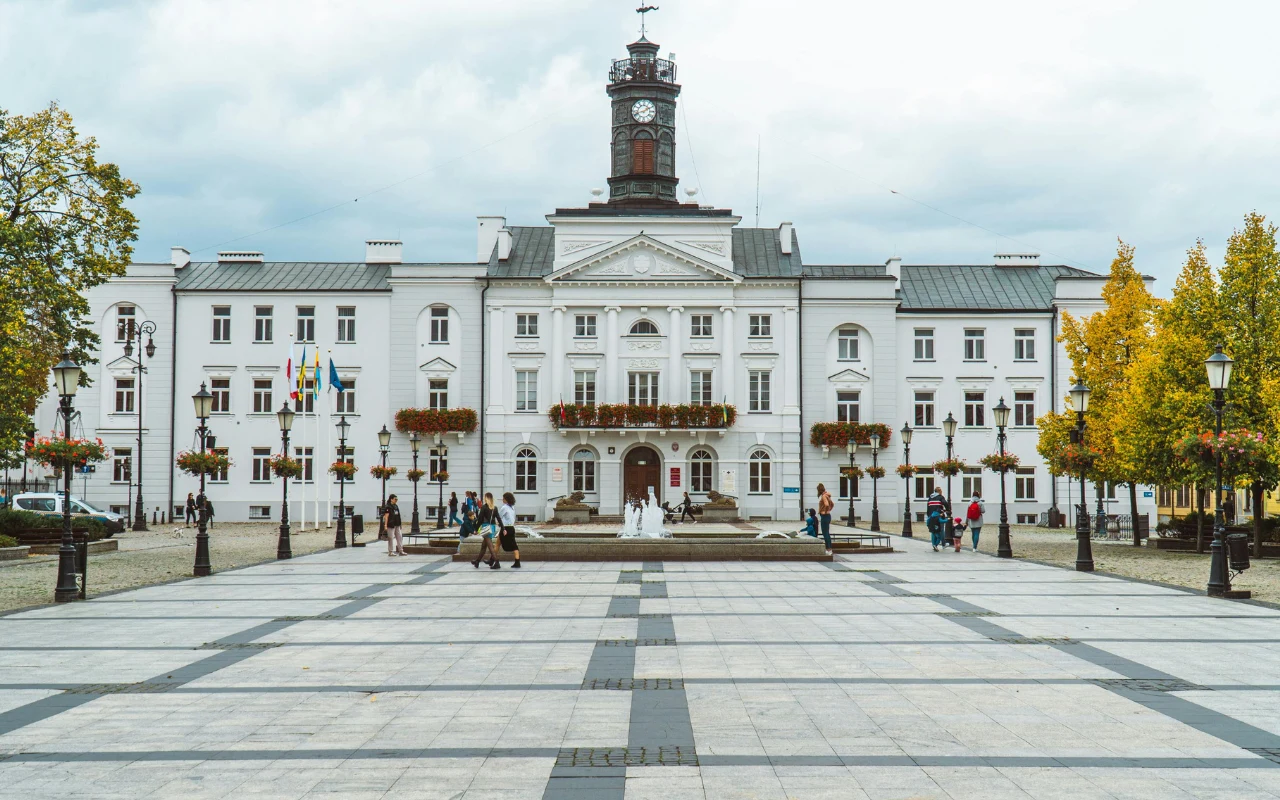 Biały budynek ratusza z charakterystyczną wieżą zegarową pośrodku szerokiego, wyłożonego kamieniem placu, otoczony nielicznymi drzewami i lampami ulicznymi, z grupami spacerujących osób.