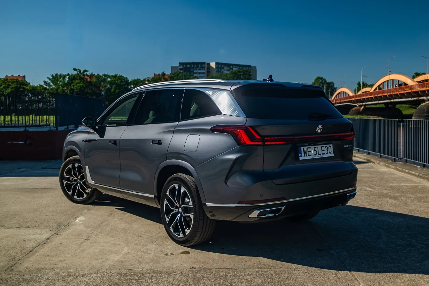 Nowoczesny, ciemnoszary SUV z charakterystycznymi tylnymi światłami LED i dużymi, czarnymi felgami stoi na otwartym placu w słoneczny dzień, w tle widoczny most łukowy oraz elementy miejskiej zabudowy.