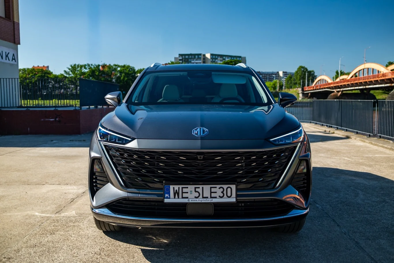 Nowoczesny SUV marki MG w kolorze grafitowym, widoczny z przodu z dużym, charakterystycznym grillem, na tle miejskiej architektury oraz mostu po prawej stronie.