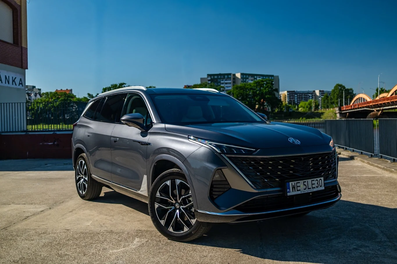 Nowoczesny, szary SUV o dynamicznych liniach nadwozia i efektownych felgach, zaparkowany na otwartym placu w słoneczny dzień, z widoczną czarną atrapą chłodnicy i nowoczesnymi reflektorami LED.