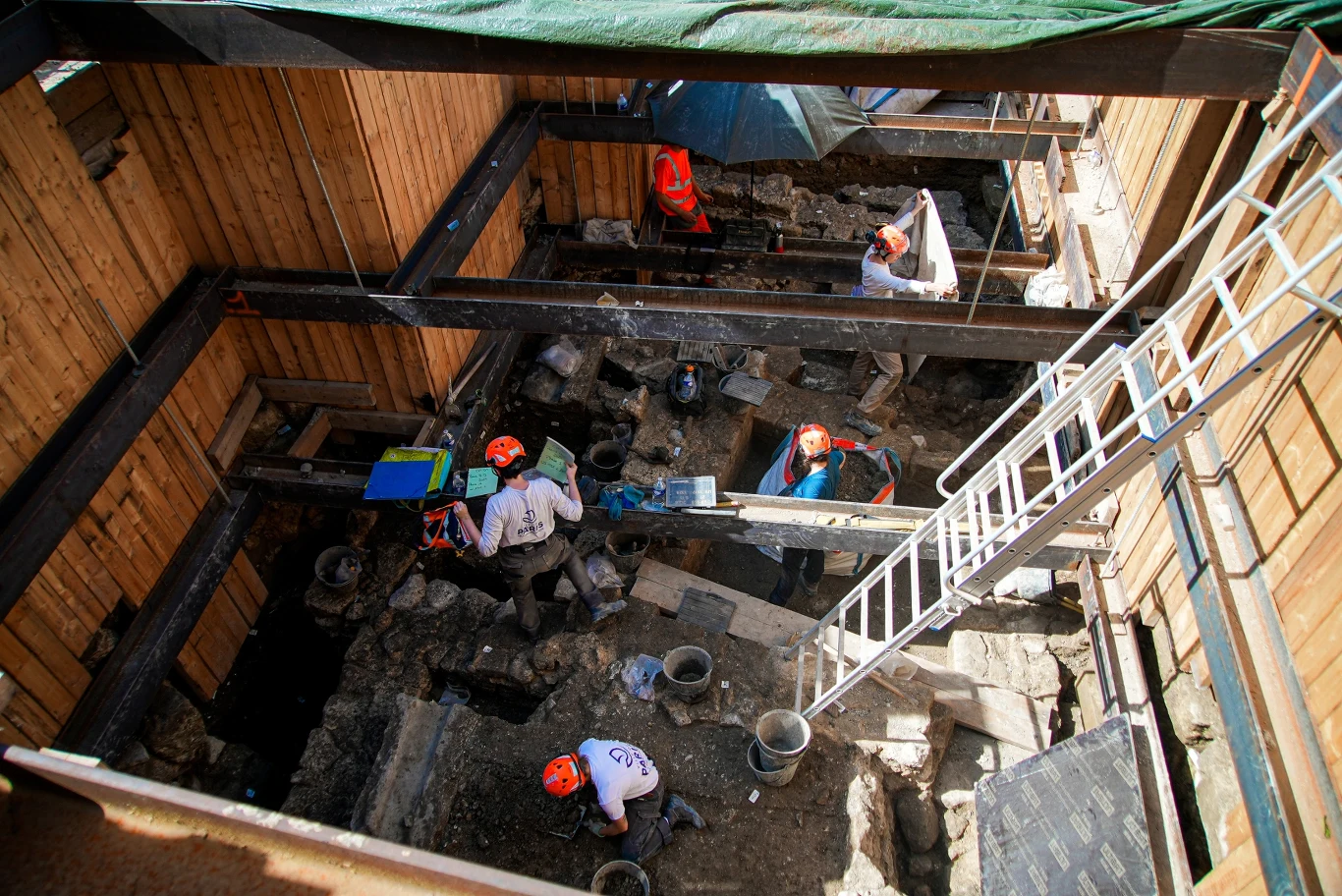 Archeolodzy kopią cztery metry pod ziemią podczas wykopalisk obok Notre Dame Grupa osób w kaskach ochronnych prowadzi prace archeologiczne w głębokim wykopie otoczonym drewnianymi ścianami, używając specjalistycznego sprzętu i narzędzi.